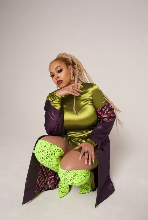 Harleighblu crouching down in a satin Chartreuse green dress, green kneehigh boots and purple long jacket with blonde dreadlocks in a high ponytail