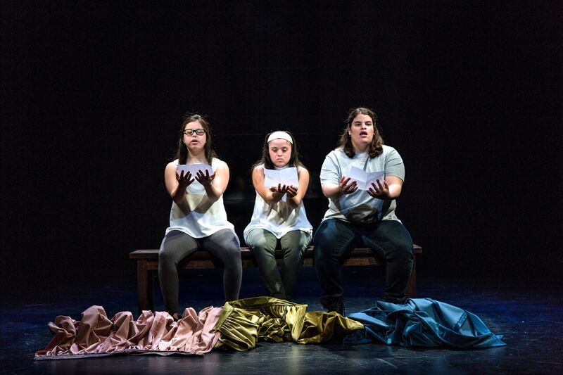 Three actors sit on a bench holding out paper to read, pink, green and blue fabric on the floor at their feet