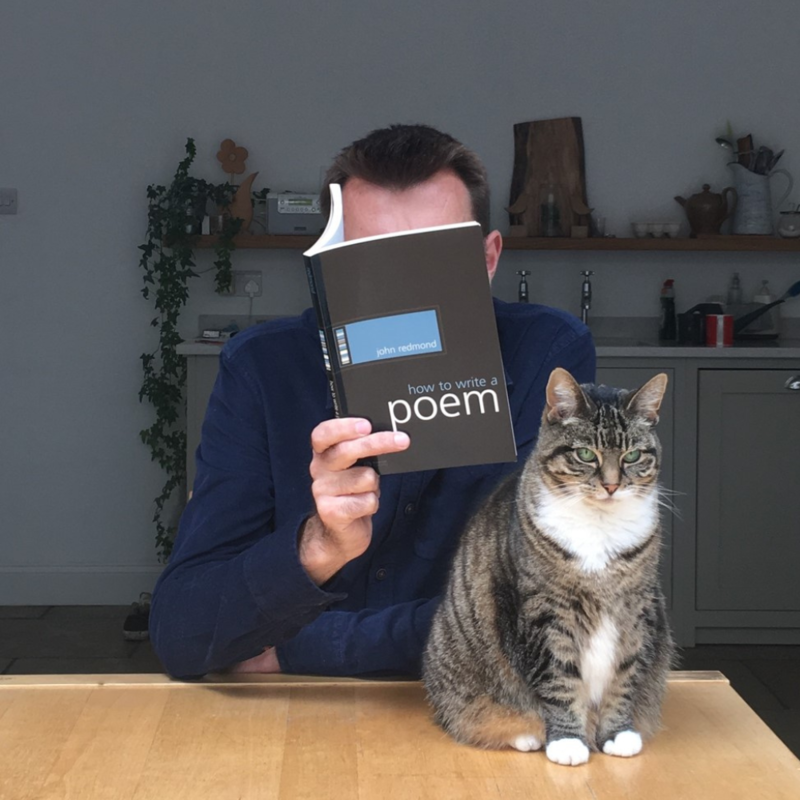 Brian Bilston sat at a table with a cat. He's reading a book which is obscuring his face