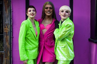 Billie Gold, Alex Fincher and Alfie Ordinary are stood in front of a bright pink building. Billie and Alfie wear bright green glittery suits, and Alex wears a bright pink coat over a bright pink bra