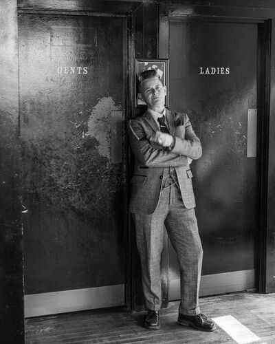 Black and white picture of Joelle Taylor standing in betwwen restroom doors which say 'Gents' and 'Ladies. Joelle is wearing a suit and has her arms crossed.