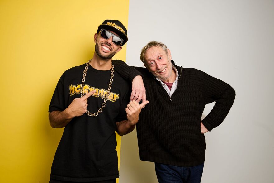 Michael Rosen and MC Grammar standing over a yellow and white background. MC Grammar is pointing at Michael Rosen, and wearing a cap, sunglasses and a gold chain. Michael Rosen has his hand on his hip smiling at the camera