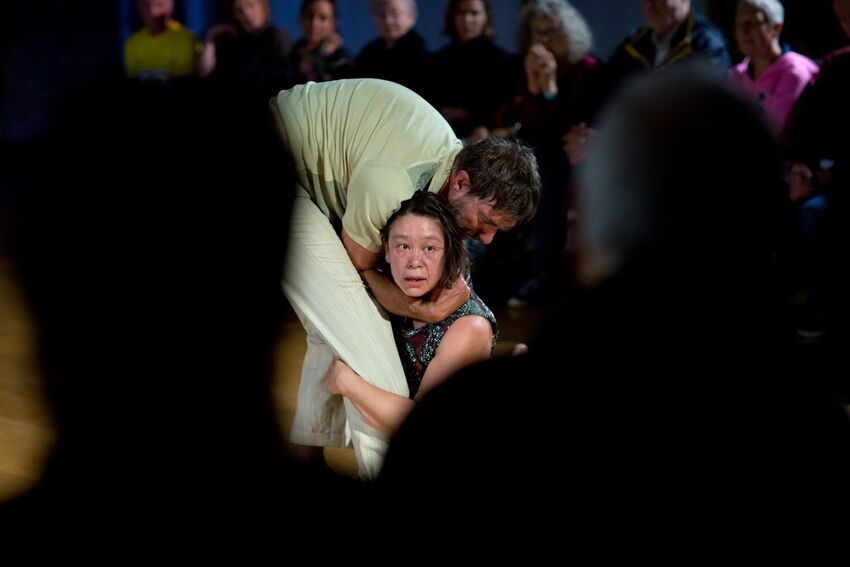 An asian woman sits on the floor and is hugged by an older white man
