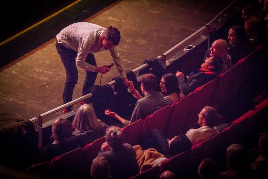 A comedian on stage bends down to interact with his front row audience