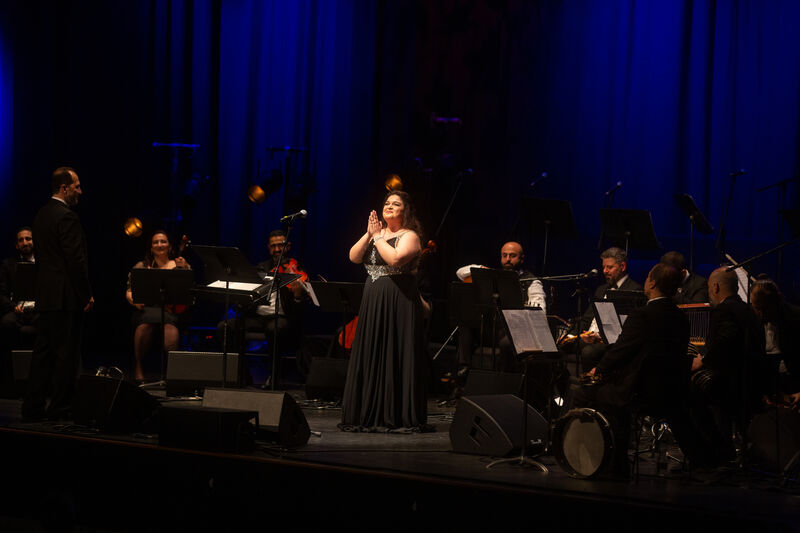 Woman in dress stands centre stage, her hands held up together, she is surrounded by musicians
