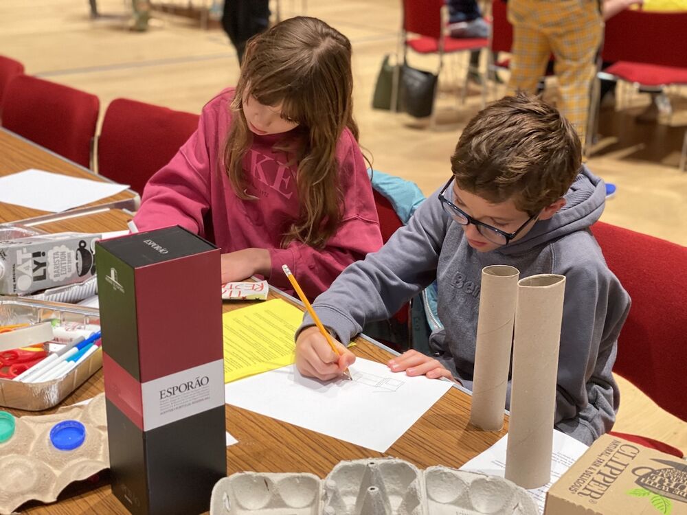 Two young children sit at tabled crafting. They are deep in concentration and surrounded by craft materials