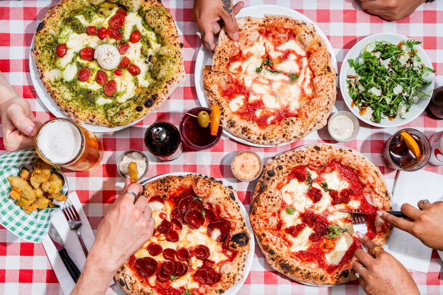 A red a white table cloth sits below a spread of four pizzas, people are reaching in to take a slice