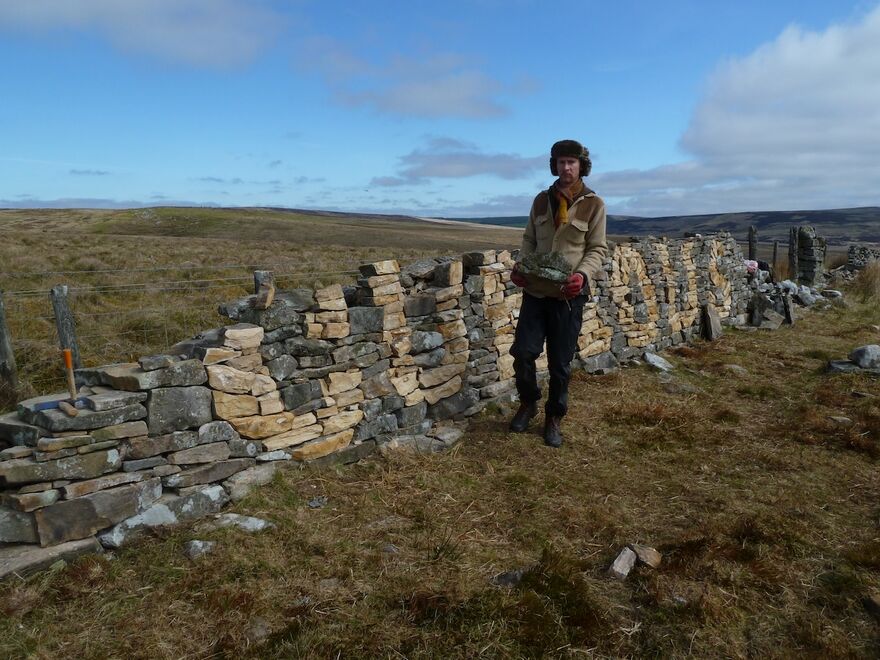A person stands next to a traditionally built slate wall. They are wrapped up against the elements and the sky is blue and cloudy