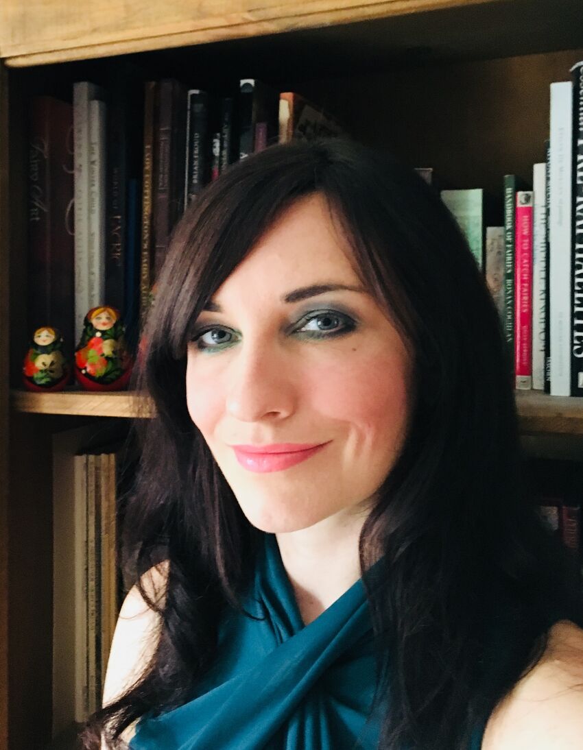 A portrait of Michelle Harrison. She has shoulder length dark brown hair, blue eyeshadow and pink lipstick. She wears a blue top. Behind her is a bookshelf full of books.
