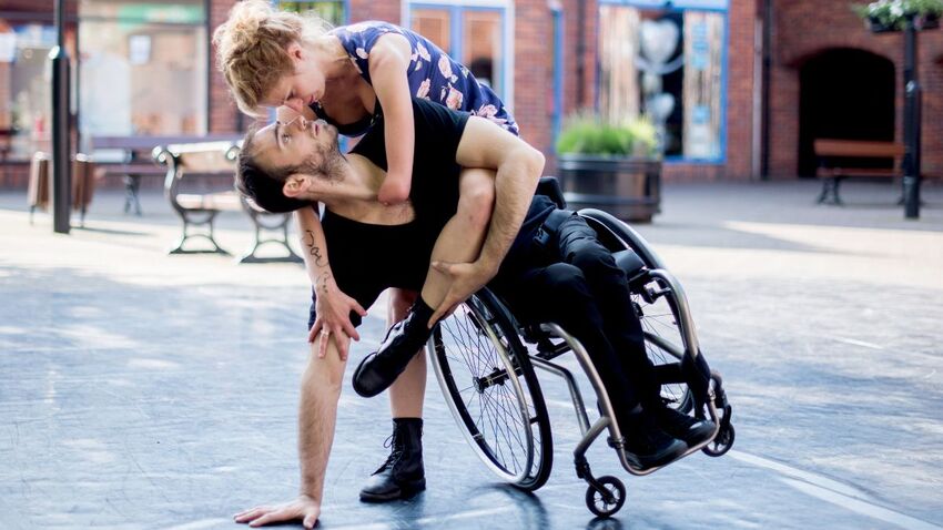 Two dancers are embracing. A male dancers is in a wheelchair which is tipped onto the edge of one wheel, and the female dancer leans over him.