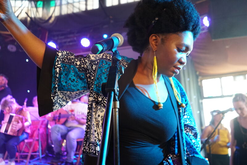 A Black woman with an afro wearing a patterned shirt and black top stares at the ground with one arm in the air. She's stood in front of a microphone stand