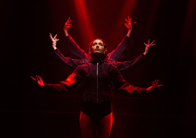 Three dancers stood one behind the other at different stages of lowering their arms, lit from above by a red light