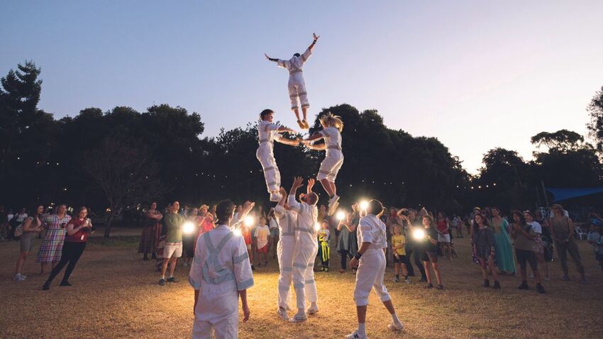 Seven acrobats dressed in white are performing a stunt. Two acrobats stand on two others shoulders, while another is thrown into the air. It is dusk, and the audience is surrounding the acrobats holding lights. 