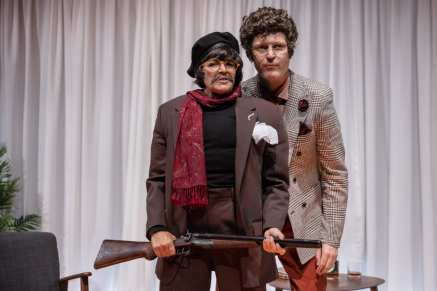 Two people stand together. One with a black beret, round yellow glasses and moustache, the other in glasses, a short curly wig and brown checked blazer