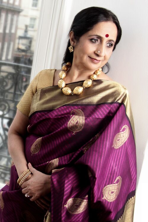 Aruna Sairam, stands against a white wall. Her hands are clasped together and she looks to her left.