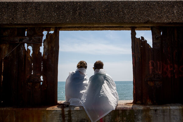 2 People in Plastic Ponchos at Brighton Festival event Last Resort, sitting together, on a wall, staring at the sea. Photo by Victor Frankowski
