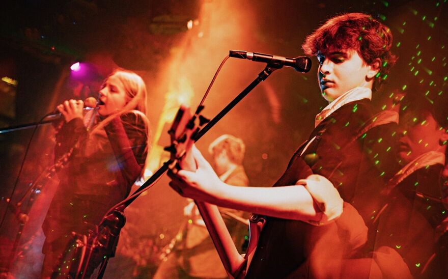 Two young people on a stage, one playing a guitar and the other singing into a microphone