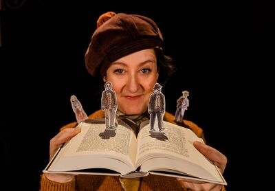 A woman in a brown beret holds an open book. Stood on the book at tiny cut out characters