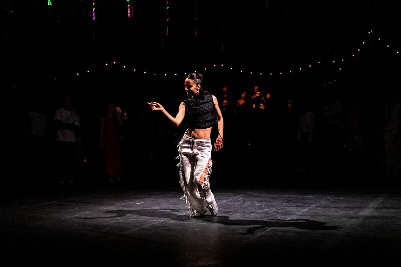 A dancer is in the middle of the dancefloor with a spotlight on them, as they point to one side of the stage