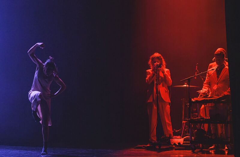 A single dancer stands on the left of the image, dancing alone on a black stage – she is lit only by a single blue light. Her left leg is touching the stage while her right leg is raised and bent at the knee, and her left arm grabs her hip while her right arm is curved above her head with her hand loosely closed. She is wearing a light-coloured dress and her dark hair is tied up in a bun. On the right of the image, separated from the dancer by an unlit area of the stage, a group of male musicians play various instruments, lit by a stark red spotlight. They are dressed in suit trousers and blazers