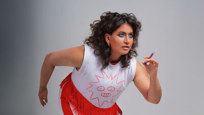 Leila Navabi in a running pose, wearing a white top and red trousers. They are holding a medicine syringe 