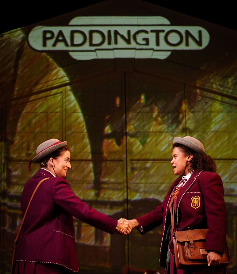 Two people wearing burgundy school uniforms shaking hands in front of a 'Paddington' station sign