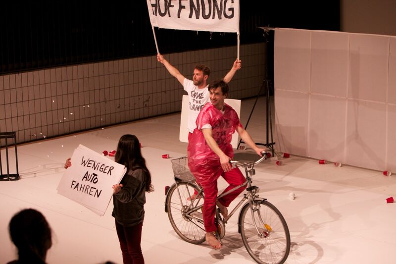 A performer wearing a red rain poncho cycles across the stage