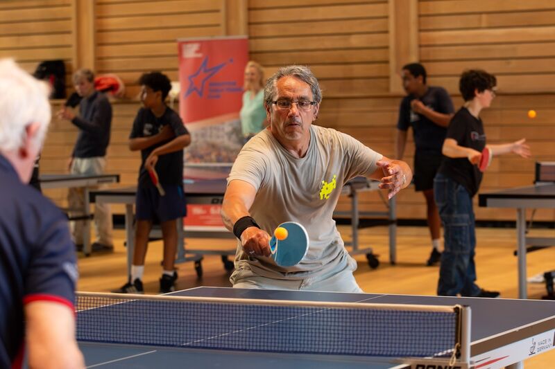 Two older men are playing table tennis. They are both concentrating but having fun. In the background people of all ages are also playing