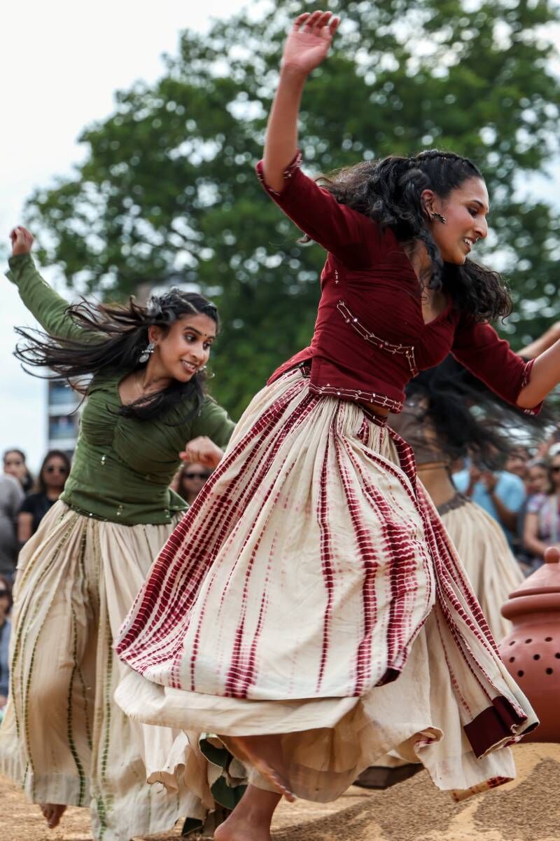 Two dancers skip around a sandy floor, waering floaty skirts and their right hand swung up in the air