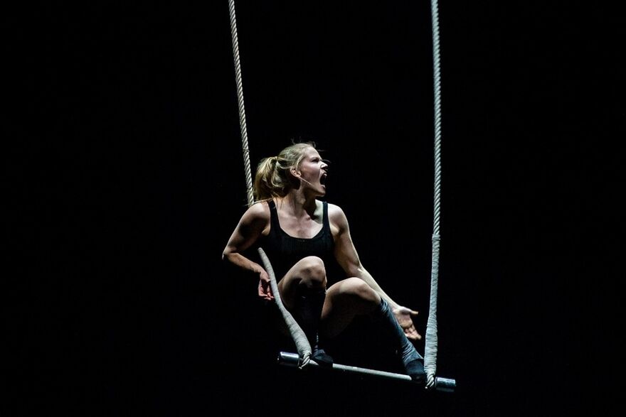 A woman balances on a trapeze, crouching with her mouth open in a shout