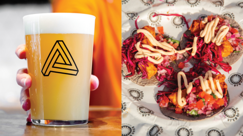 A close up of a hand holding a pint of beer next to a plate of three tacos