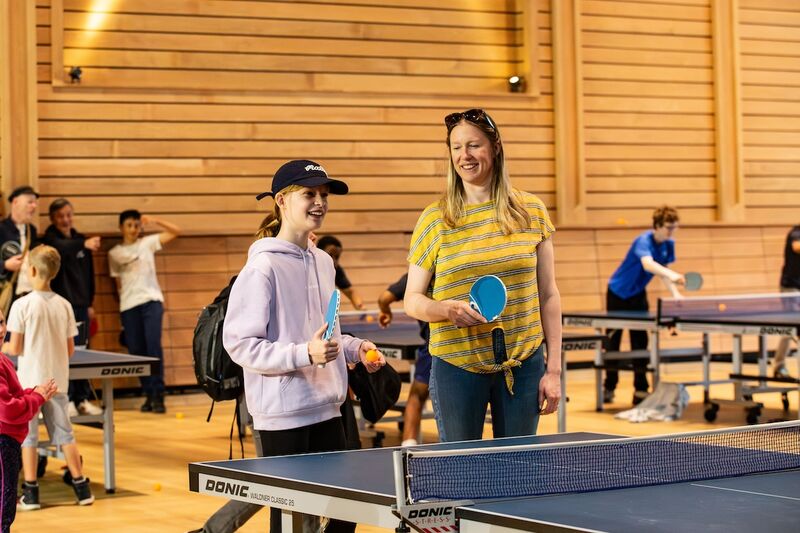 A mother and daughter are stood at one end of a table tennis table holding bats and the ball. They are both enjoying themselves and having fun