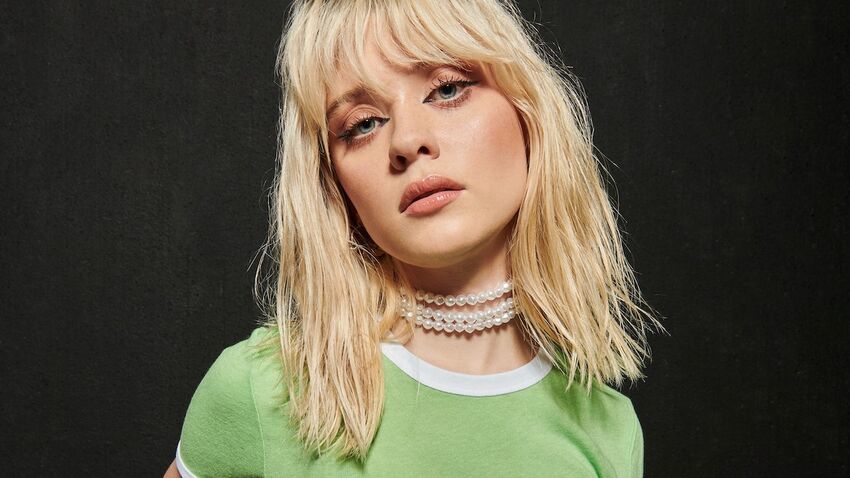 Maisie Peters wearing a green t-shirt and a white beaded necklace
