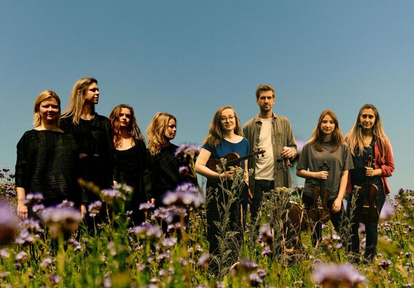 Four musicians holding violins standing in a field with purple flowers. 