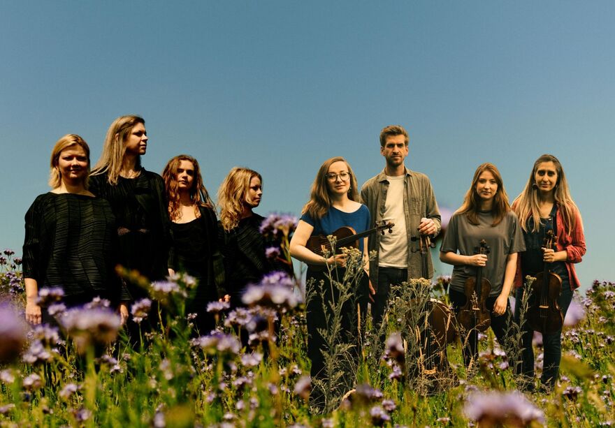 Four musicians holding violins standing in a field with purple flowers. 