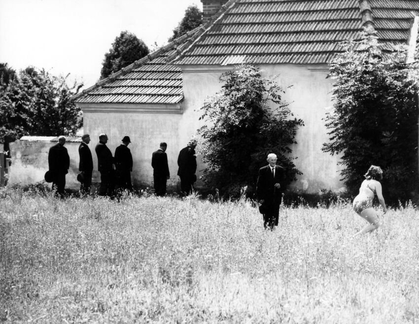 Greyscale photo of men in black suits standing outside of a country house with a woman in her underwear