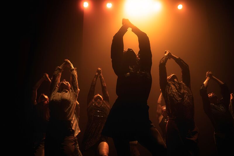 Group of dancers stand upright together, their heads pointed towards the ceiling and their hands clasped together, held high above their heads. There is an orange light above the dancers, but the dancers themselves are shrouded in shadows. They are all dressed in dark-coloured shirts.