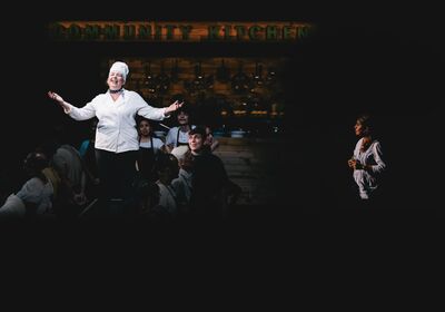 A woman in a chefs costume sings on stage, with people in cooking aprons around her, a wall of pots and pans behind them