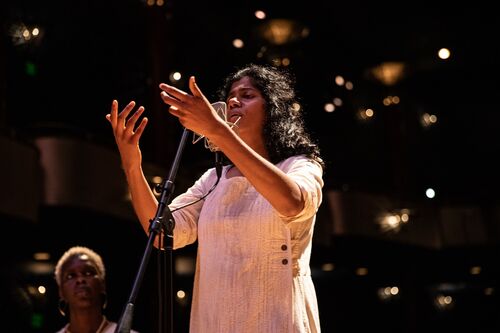 Ganavya sings into a microphone, her arms raised up, palms open