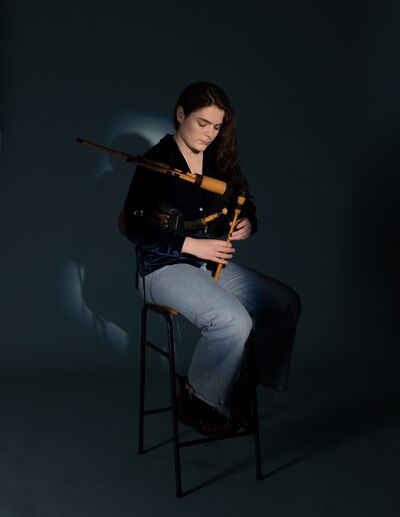 A young woman wth long black hair, swept to one side, sits on a stool holding her 'small pipes' instrument. She wears a black top and grey trousers.