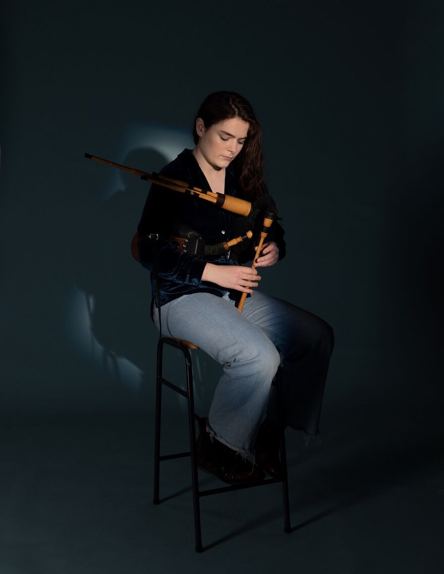 A young woman wth long black hair, swept to one side, sits on a stool holding her 'small pipes' instrument. She wears a black top and grey trousers.