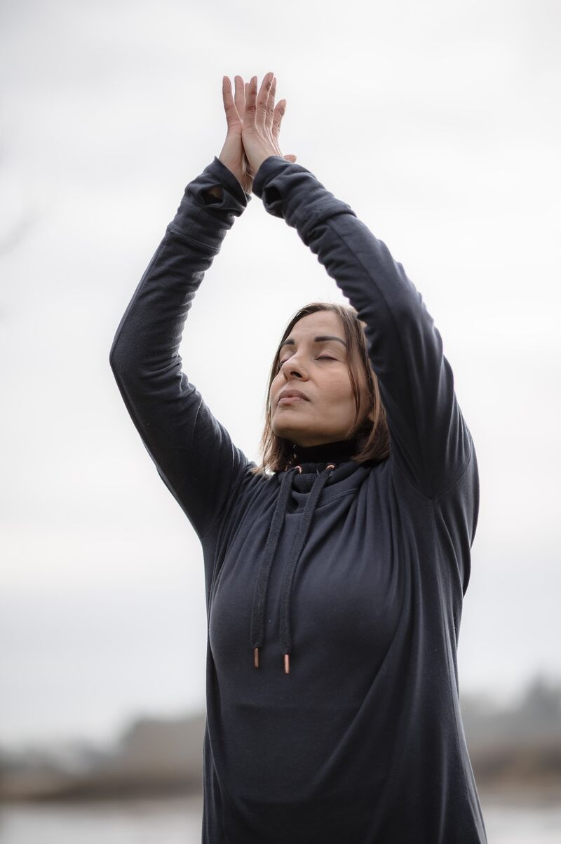A woman stands outdoors. Her arms reach into the air with her palms together. Her eyes are closed and she has a calm expression on her face