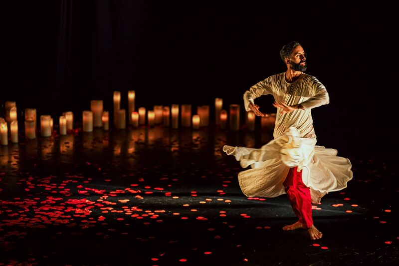A man in a white dress with red trousers underneath dances on a black stage with red petals on it, behind him is a row of white candles.