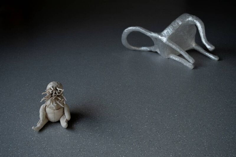 A small alien clay creature sits on the floor in front of a miniature chair made of cling film lay on its side,