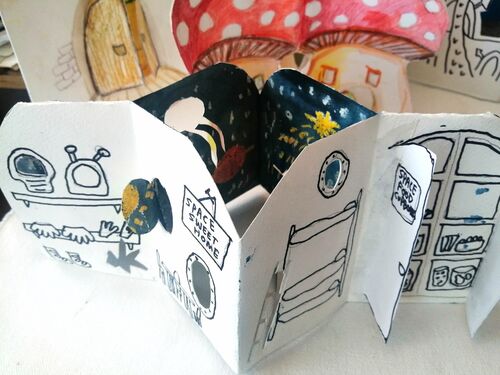A paper craft dream home stands on a table. It is space ship themed and has portholes and doors that open
