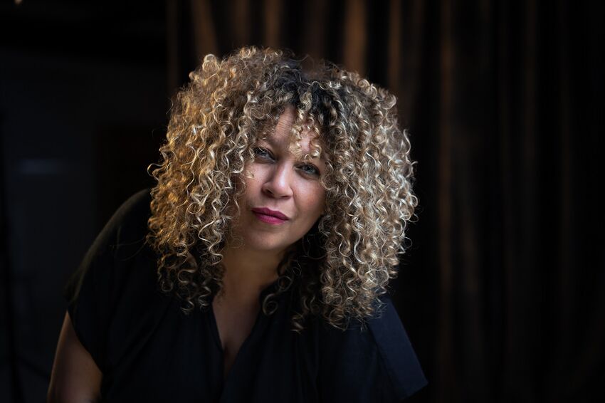 A Jamaican-mixed heritage woman with curly hair leans forward slightly, a small smile on her face