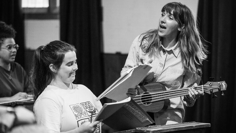 A girl sits at a desk holding a notebook while a woman next to her sings and plays ukulele