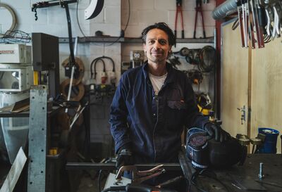 Will Nash stands in blue overalls, he's in his workshop with tools hanging on walls and on his worktop