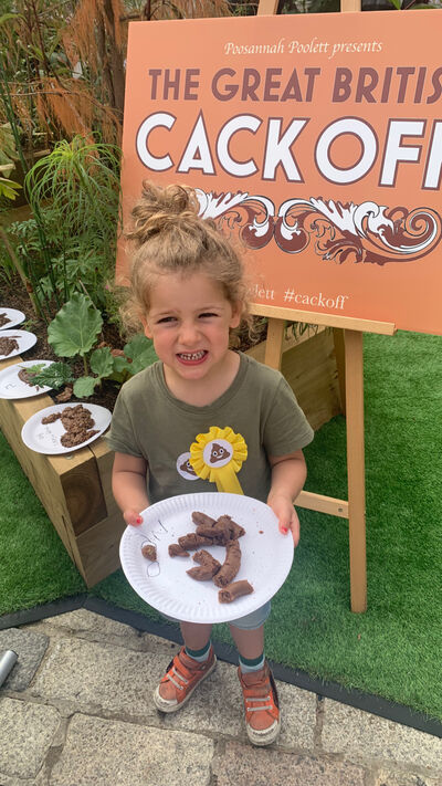 A young child holds a plate of, what appears to be poo, in front of a sign reading The Great British Cack Off