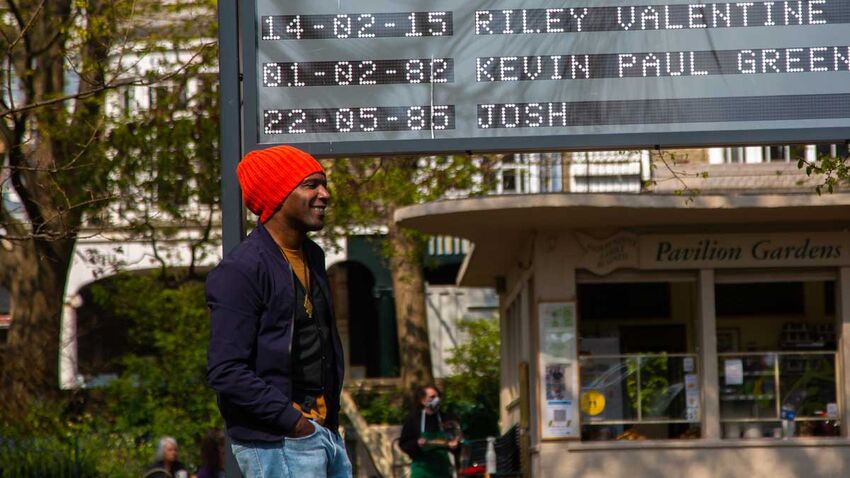 Brighton Festival Guest Director Lemn Sissay stands below the Arrivals and Departures installation board in Pavilion Gardens